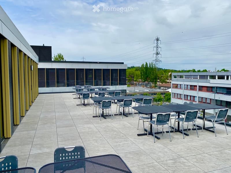 Roof terrace with tiled floor, multiple black tables and blue chairs arranged for seating, overlooking a cityscape with buildings and power lines.
