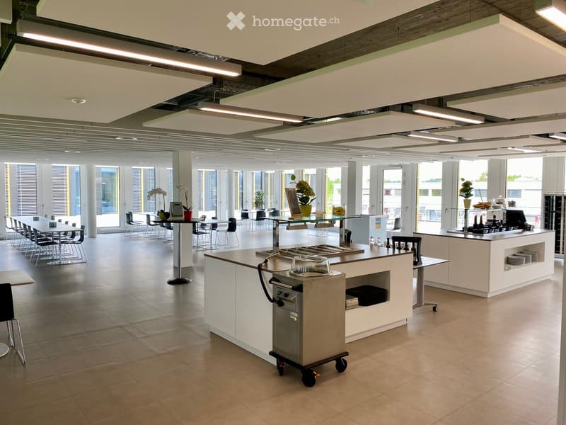 Modern kitchen area with white cabinets, stainless steel appliances, and a central island. There are multiple tables and chairs arranged for seating, along with a refrigerator, oven, and a display screen.