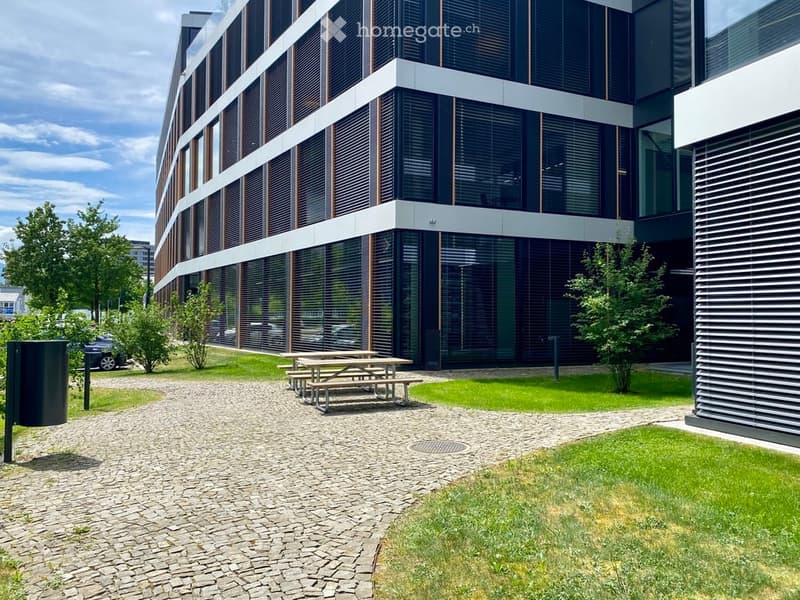 Modern multi-story building with large glass windows and a sleek design, surrounded by a paved area with picnic tables and greenery.