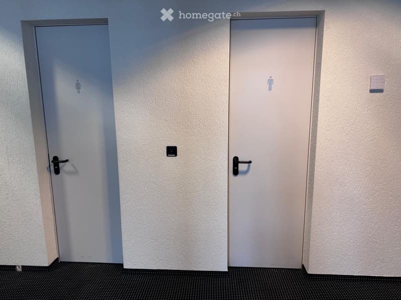 A pair of white doors with black handles. The left door is labeled 'Woman' and the right door is labeled 'Man'. The doors are set into a textured white wall, with the floor in front covered in a black speckled material.