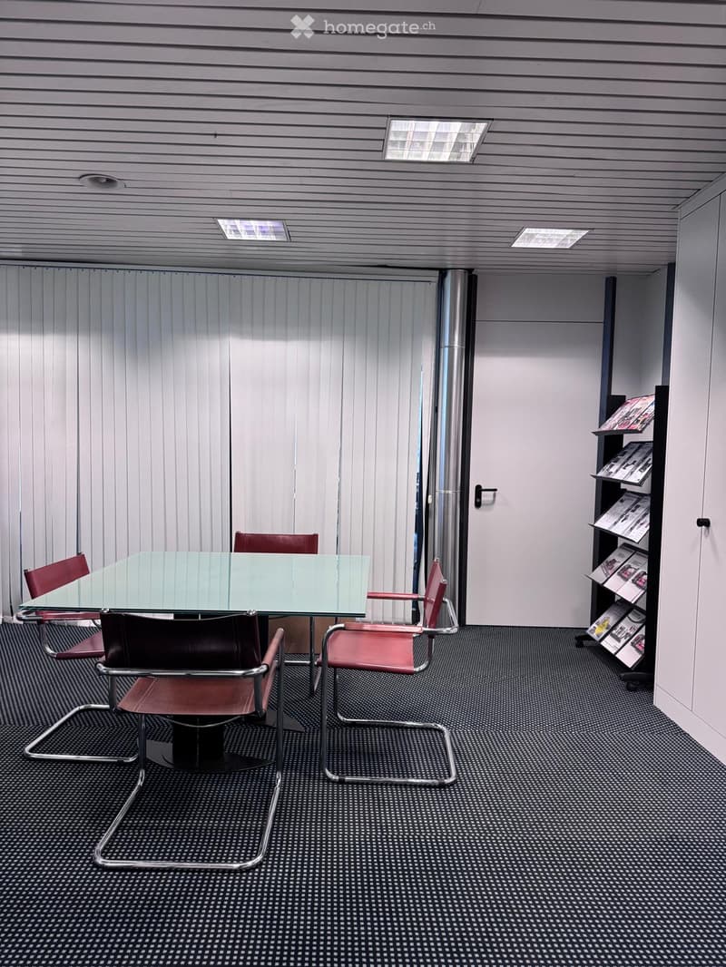 An empty room with a glass table and four red chairs with chrome base. The walls are covered with blinds and there's a door on the right side.