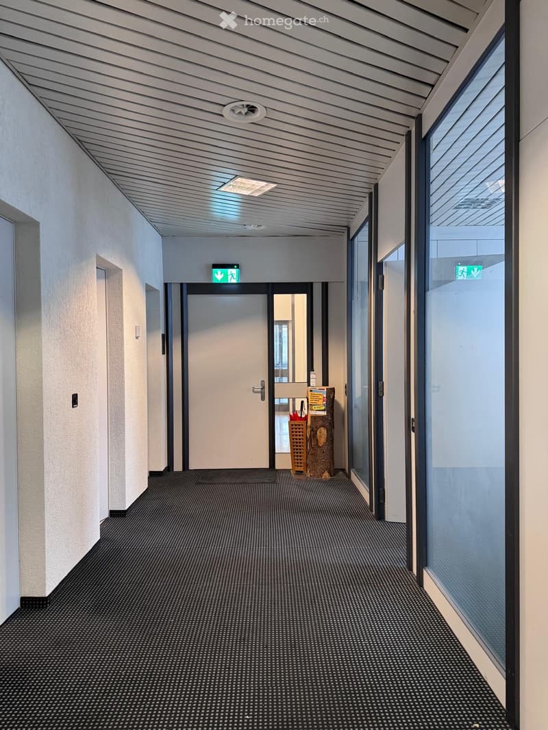 A long hallway with a white ceiling, black carpeted floor, glass doors, and white walls. At the end, there is an open white door with a black mat. There is a trash bin with a box on top next to the open door. Green exit signs are above the door.
