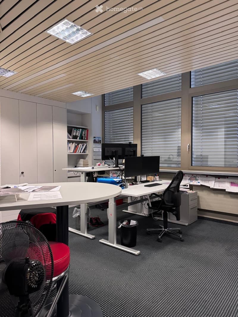 A modern office space with white walls and ceiling, two desks, office chairs, a bookshelf filled with books and documents, multiple computer monitors, a storage cabinet, ceiling lights, and large windows with blinds.