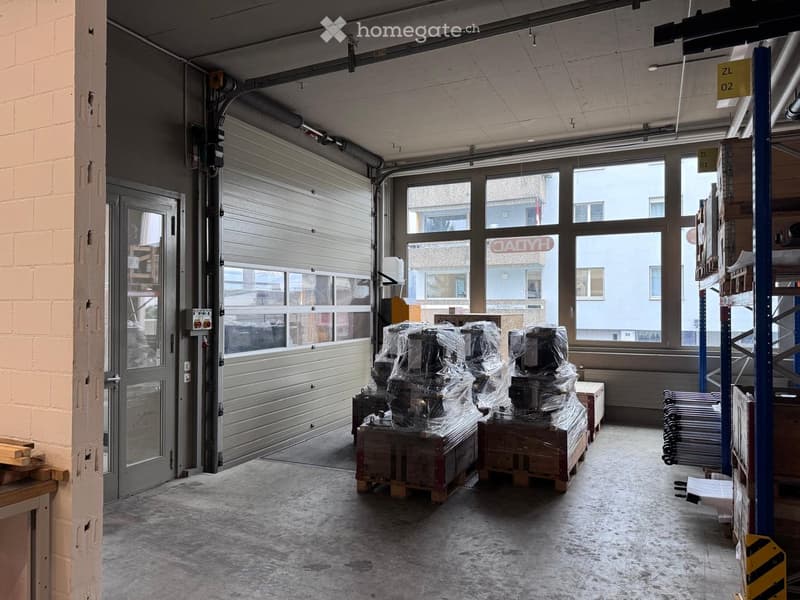 Empty warehouse room, gray door, large windows, white floor, stacks of wooden crates with items in plastic covers, storage shelves on the right.