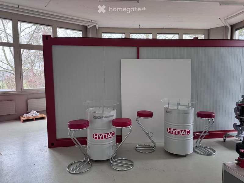 a room with a white wall, gray walls, glass barrel tables with red stools, large windows, a pile of stones on a wooden pallet, and a machine on wheels on the right side