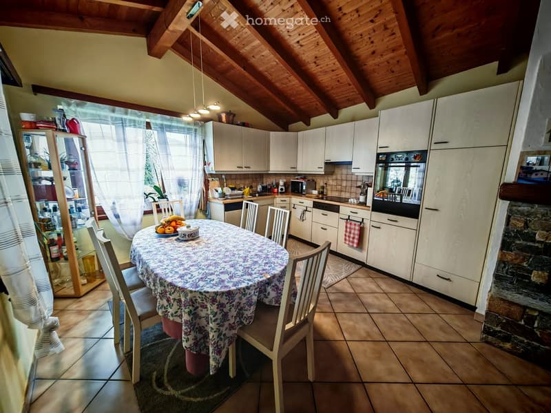 Dining table with floral tablecloth, white cabinets, tiled floor, hanging lamps, open windows, countertop with various items, shelf with bottles, and a brick column