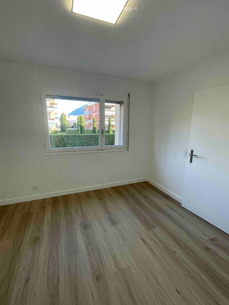 empty room with large window, wooden floors, white walls, view of a garden and buildings