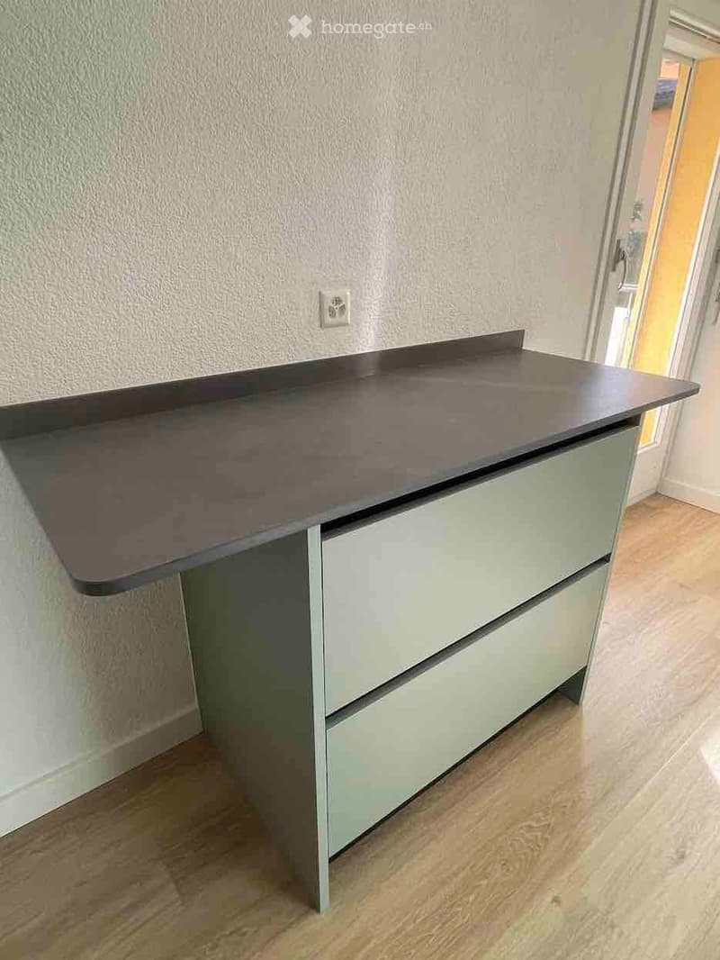Desk with drawers under, grey countertop, white and green sides, wooden floor, wall outlet