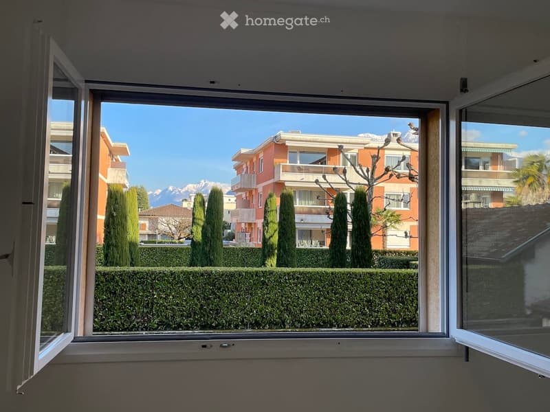Open window, view of garden with bushes, hedges and trees, orange buildings in the background, mountains