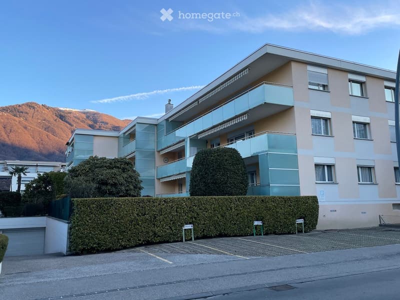 Three-story building, blue and beige facade, multiple balconies, garage, parking area, mountain in the background