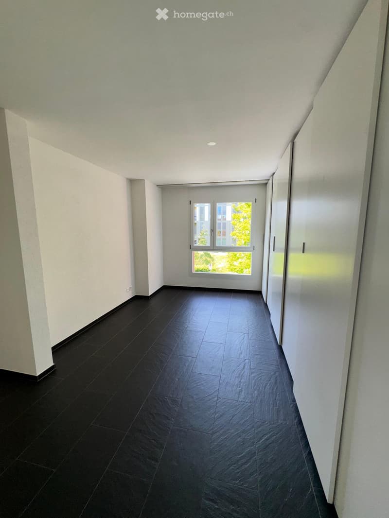 Empty hallway with dark tiled floor, white walls, and a large window at the end showing greenery outside.