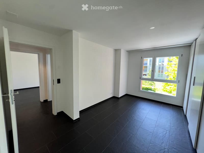 Empty room with dark tiled flooring, white walls, and large windows offering a view of greenery outside. The room features an open doorway leading to another space.