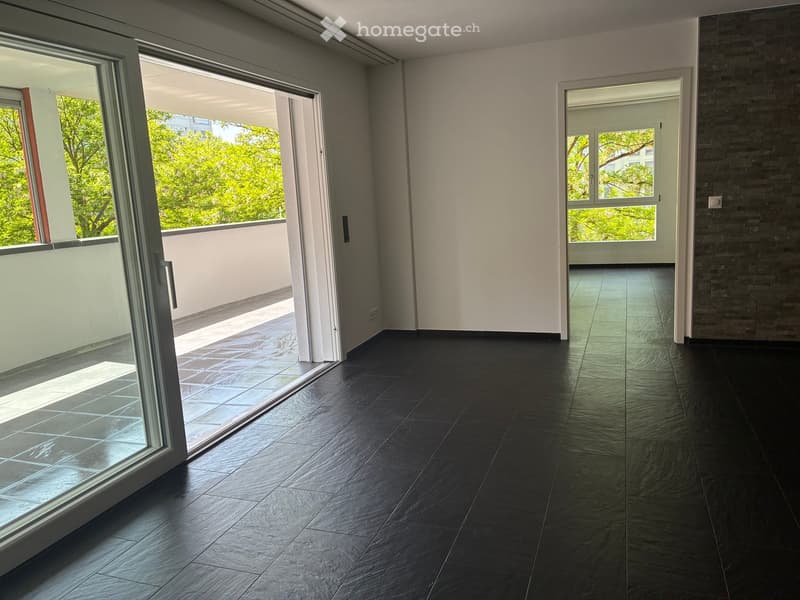 Empty room with dark tiled flooring, large glass sliding doors leading to a balcony, and a view of greenery outside.