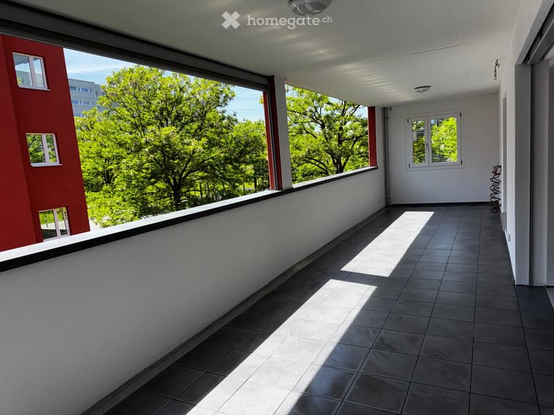 Balcony with tiled floor, white walls, and large windows offering a view of green trees and a neighboring building.