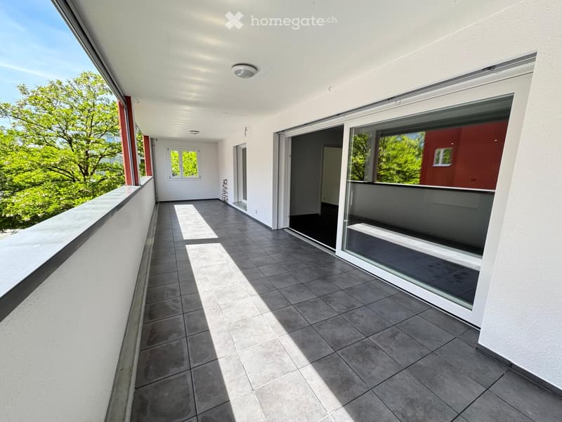 Modern balcony with tiled flooring, large windows, and sliding glass doors leading to an interior space. The balcony features white walls, a ceiling light fixture, and offers a view of greenery outside.