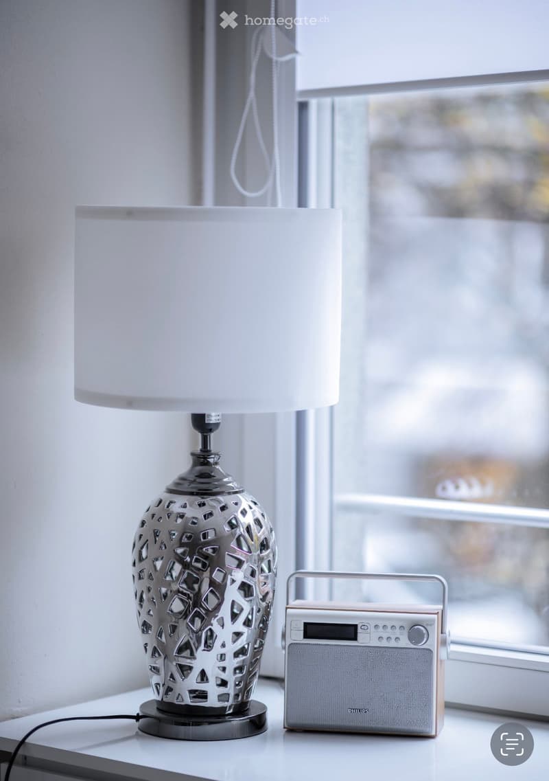 A room with a white table, lamp, and radio on the table in front of a window