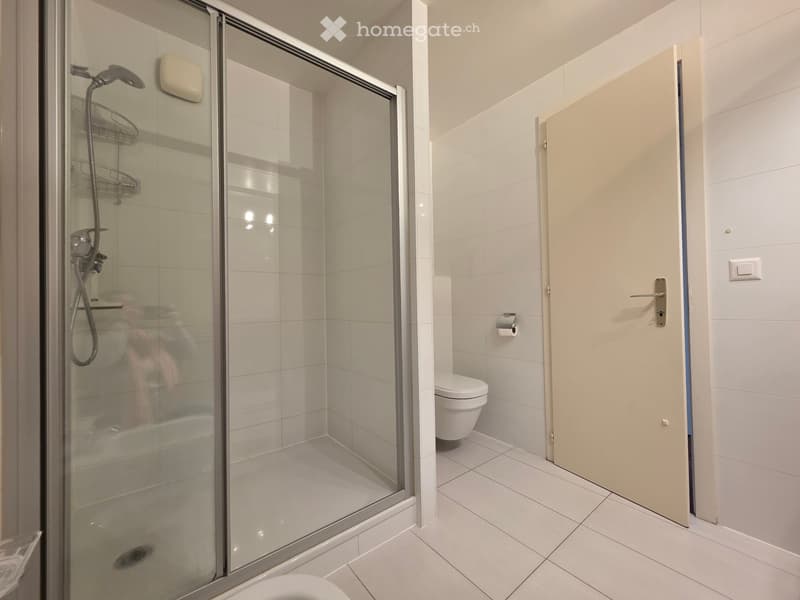 White tiled bathroom with shower, toilet, white wall, open door