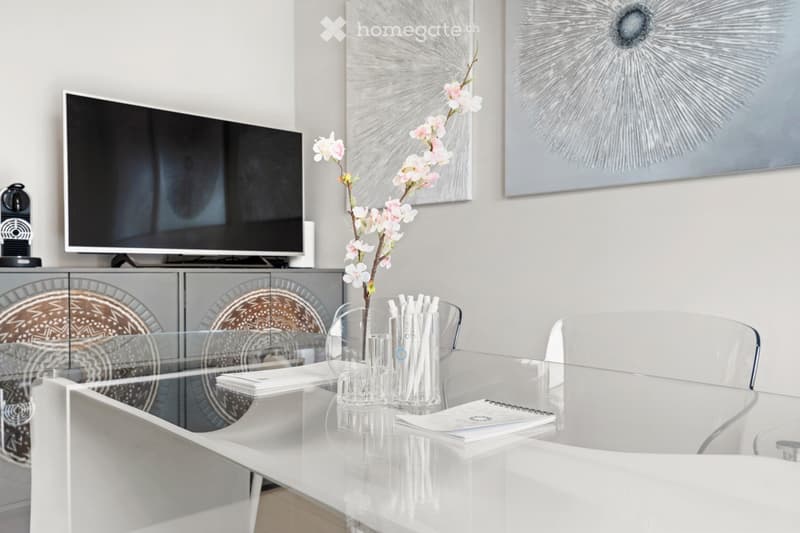 Modern room with a glass desk, clear plastic chairs, a TV, a cabinet with a glass door, a vase of flowers, and a notebook on the table
