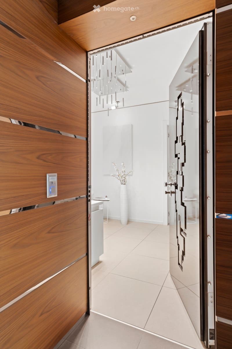 Entrance with wooden paneling, steel door with a geometric pattern, and a decorative white vase with flowers.