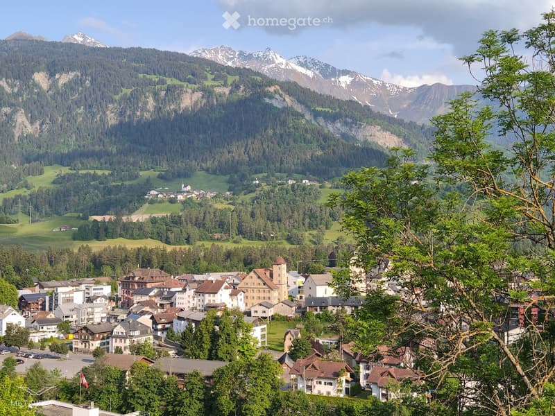 Mountain range, snow-capped peaks, town with buildings, green valley, trees, cloudy sky