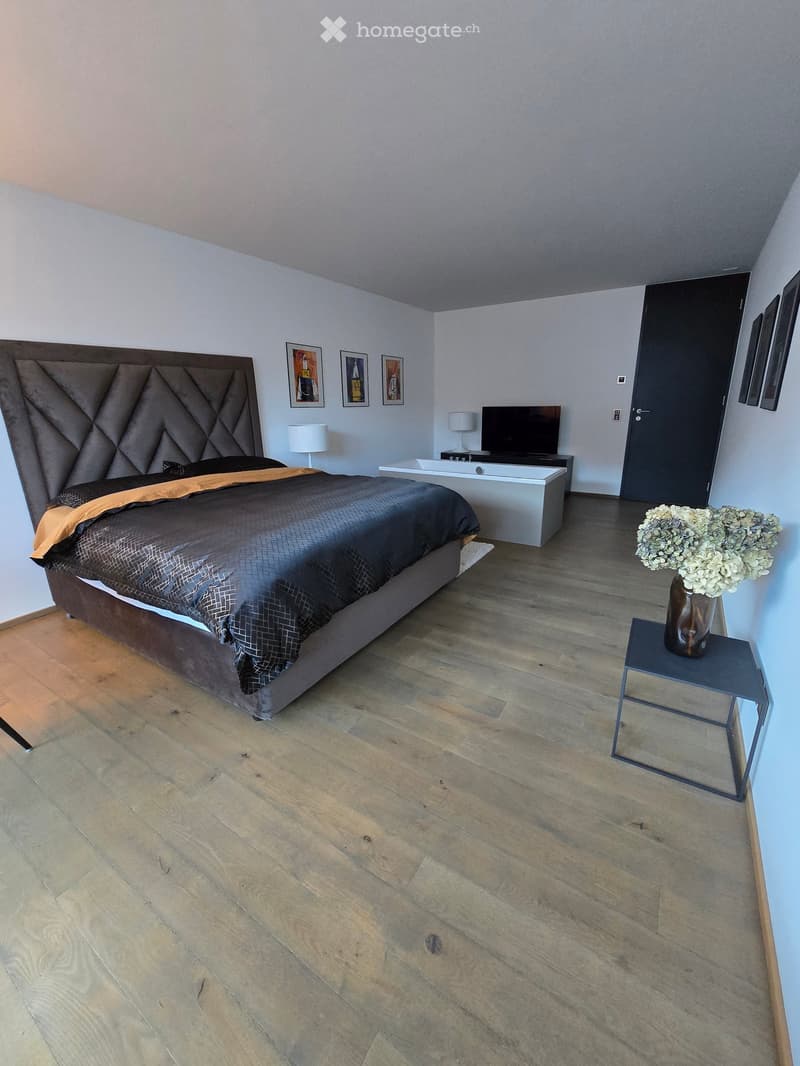 bedroom with wooden floor, bed, white walls, lamp, bathtub, flat screen TV, flowers in vase, entrance door