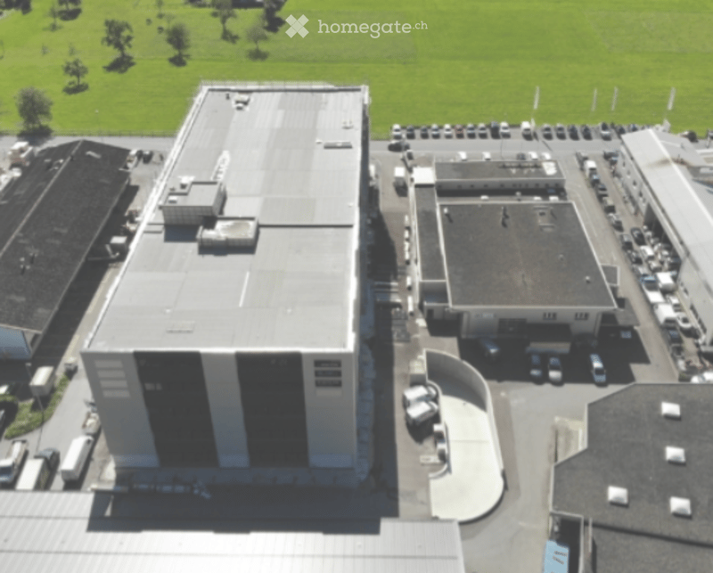 Industrial building, grey roof, multiple floors, grey wall, many cars parked, large green field in the background