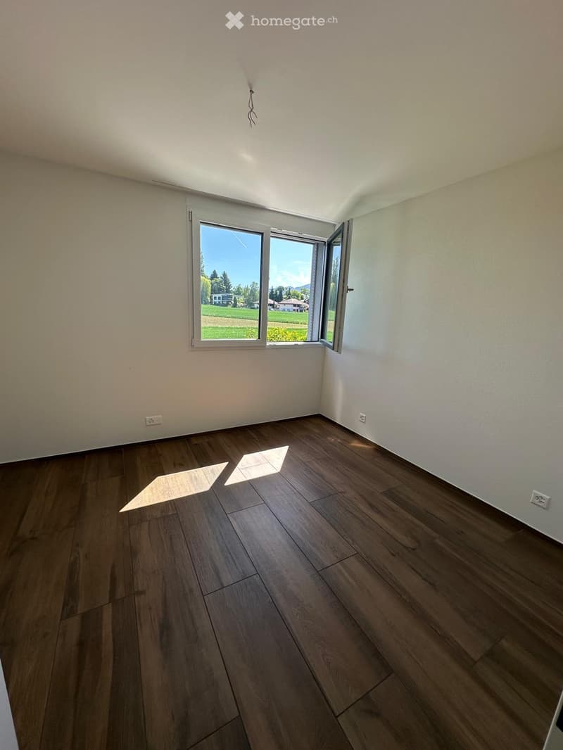 Empty room with wooden flooring and white walls, featuring a large window with a view of a green landscape.