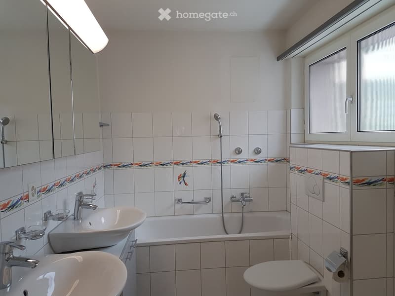 Bathroom with white tiles, bathtub, two sinks, toilet, shower head, faucets, mirrors, and a window.