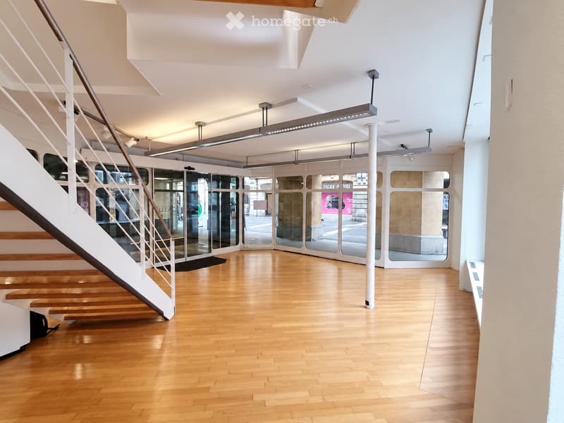 Modern, spacious hallway with wooden flooring, white walls, and a staircase leading upwards. Features large glass doors and modern lighting fixtures.
