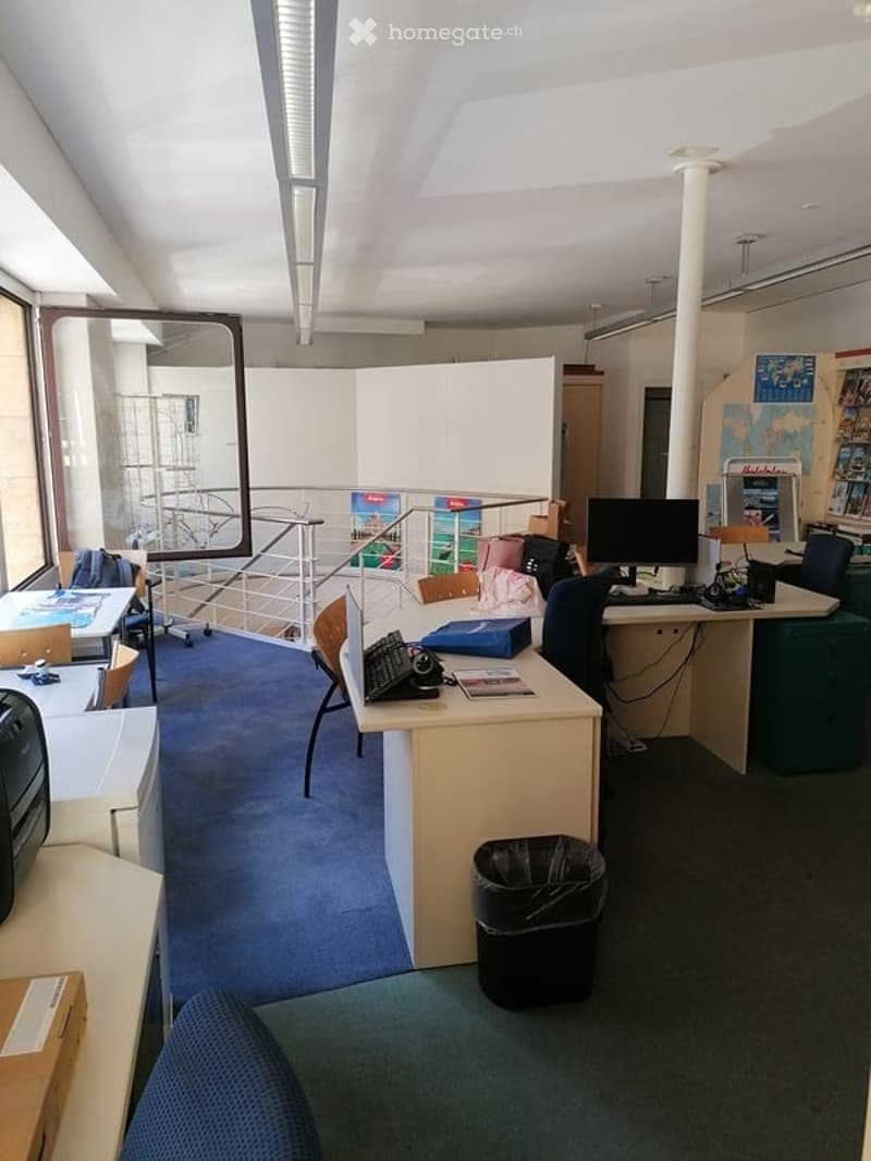 An office with a few desks, computer monitors, chairs, lamps, trash cans, and other items.