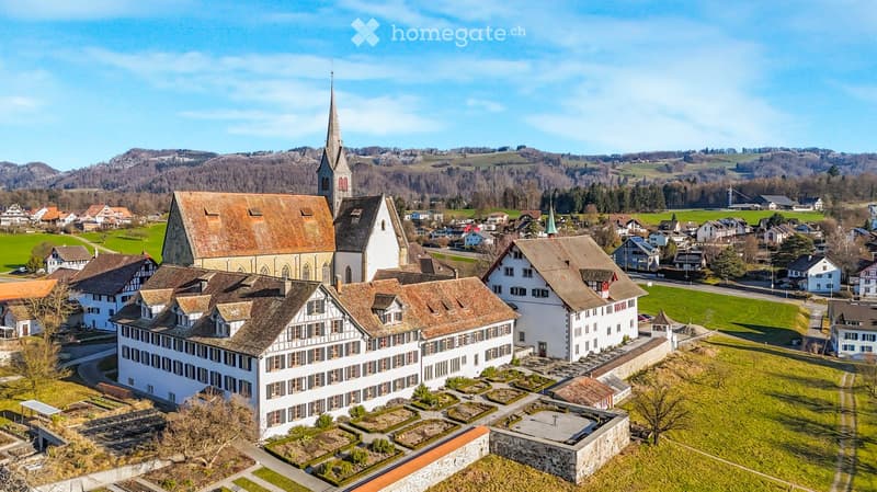 Large monastery complex, multiple buildings with varying rooftops, surrounded by green landscape, central tower, well-manicured gardens