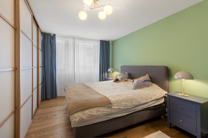 A bedroom with a double bed, nightstands with lamps, wardrobe, bedsheets, pillows, carpet, windows with white curtains, and a ceiling lamp.