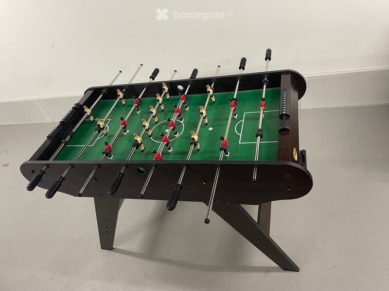 Large foosball table with a green playing surface and black metal frame, placed in a room with white walls.