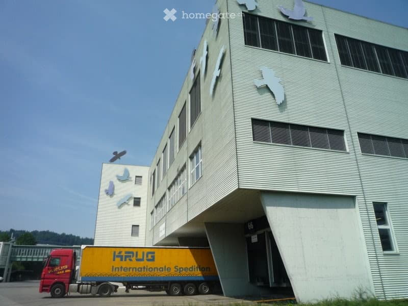 A modern architectural building with multiple windows and metallic panels, with a yellow truck parked in front of the building.