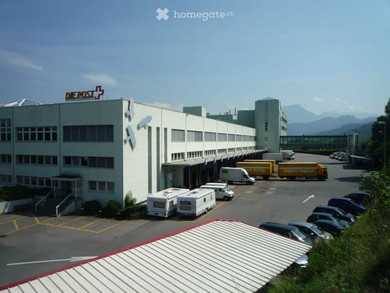 Postal facility with large parking area, several delivery trucks parked, modern white exterior, mountains in the background