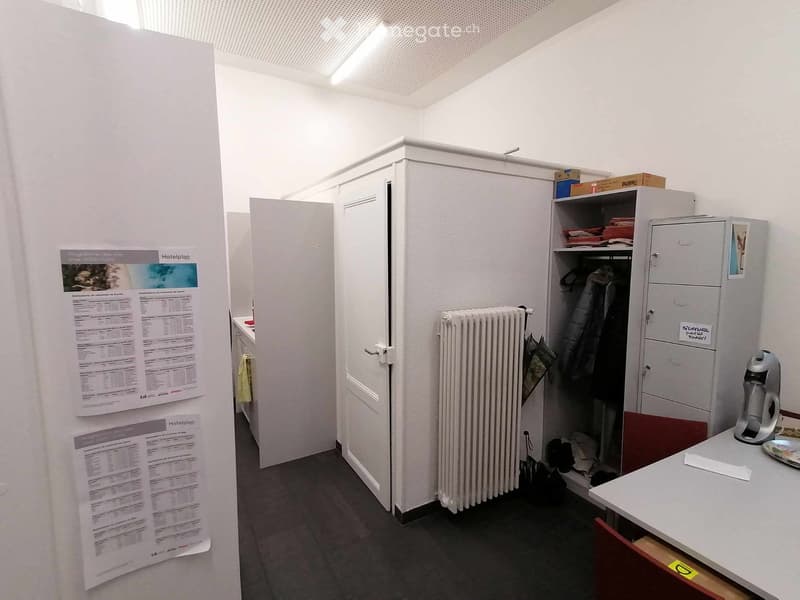Empty room with white walls, gray floor, white cabinets, radiator, papers, a table, and a cabinet with a storage box