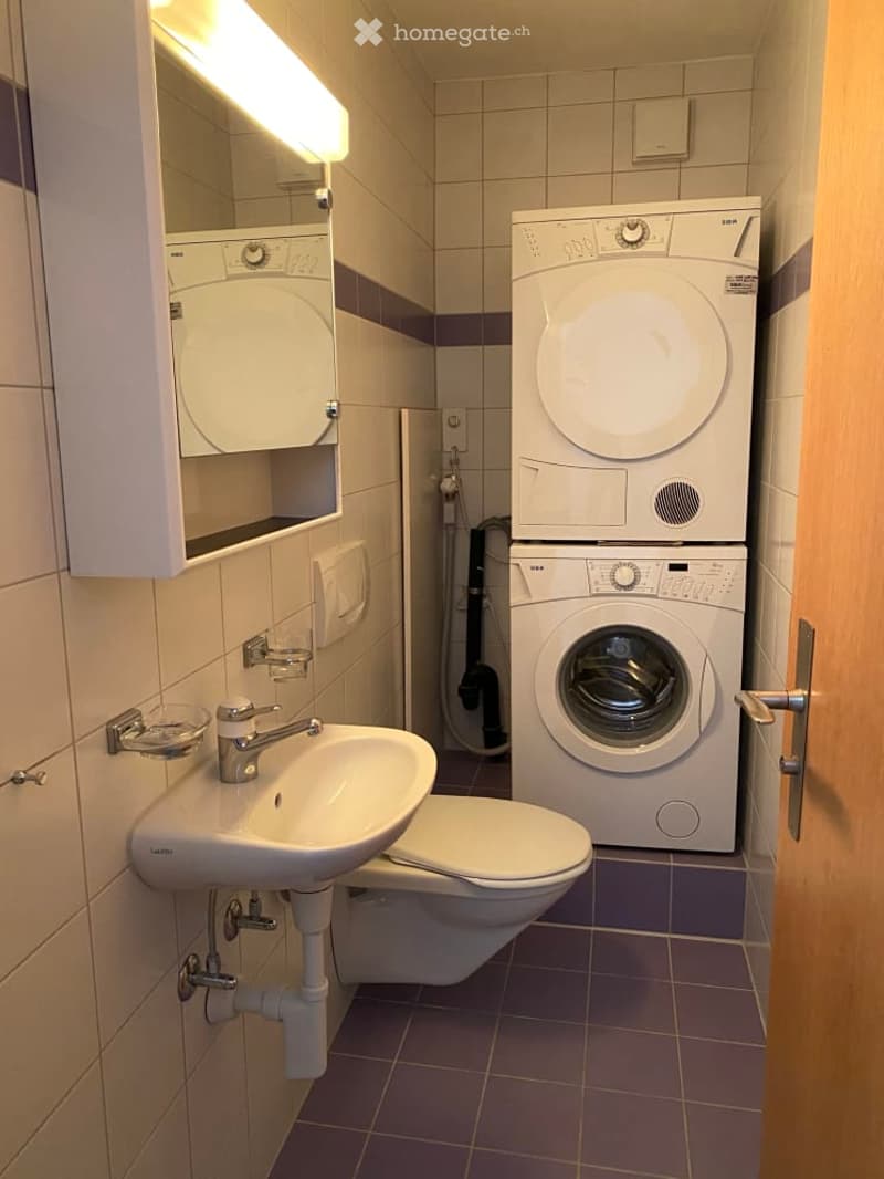 Small bathroom with white sink, white toilet, stackable washer and dryer, wall-mounted mirror, white tiled walls and purple tiles on the floor