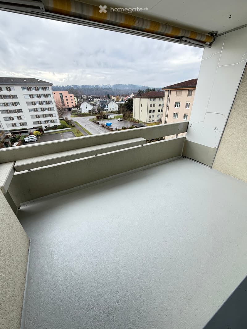 Balcony with a grey floor, a grey wall, and a metal railing. View of the parking lot and several buildings.