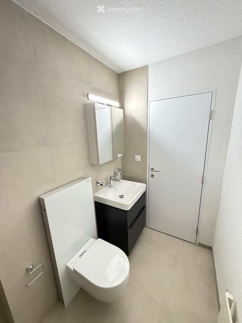 White tiles on the walls, white sink with faucet, white toilet, a white cabinet, a mirror above the sink, and a white door.