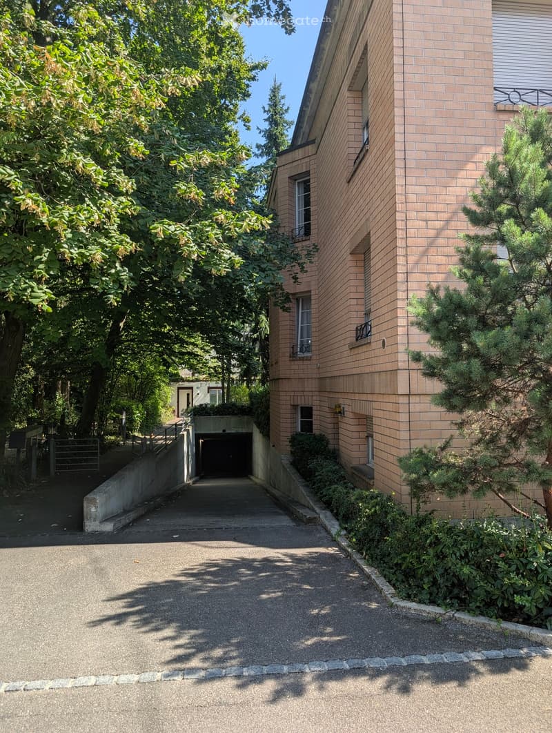 Multi-story building with brick exterior, a concrete ramp leading to the garage entrance, windows with shutters, greenery including trees and shrubs, and a paved parking area.