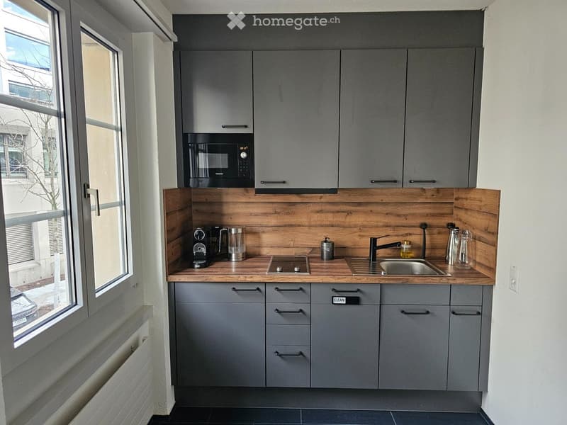 Modern kitchen with wooden countertops, grey cabinets, appliances including a microwave, coffee machine, sink, stove and dishwasher, window, radiator
