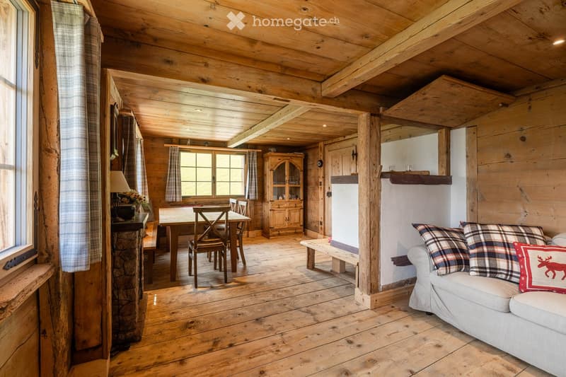 Cozy wooden interior with exposed beams, a sofa with plaid pillows, a dining table with chairs, a wooden cabinet, and a bench near the window.