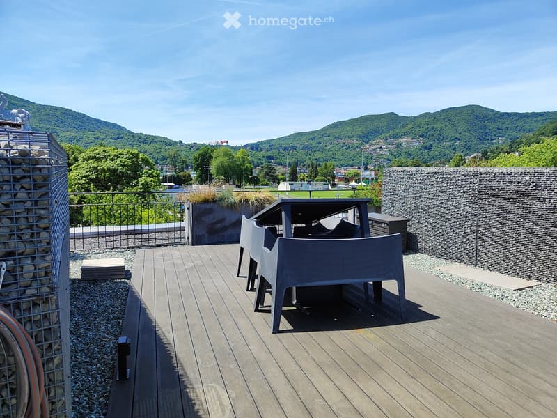 Modern wooden deck terrace with a view of a city and mountains, featuring a grey chair, a table, and a stone wall.