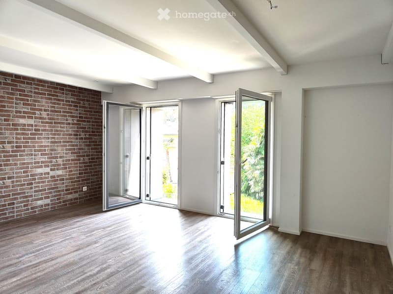 An unfurnished room with wooden flooring, white walls, and large sliding glass doors leading to an outdoor area. The room features a brick accent wall on one side and ample natural light.