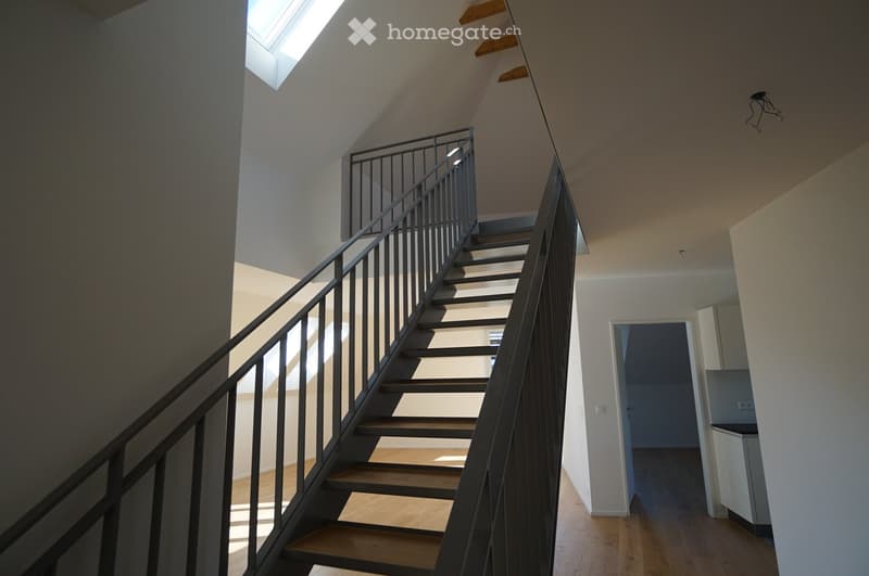 Interior view of a modern building featuring a sleek staircase with dark grey handrails, wooden steps, and an open, airy layout illuminated by natural light.