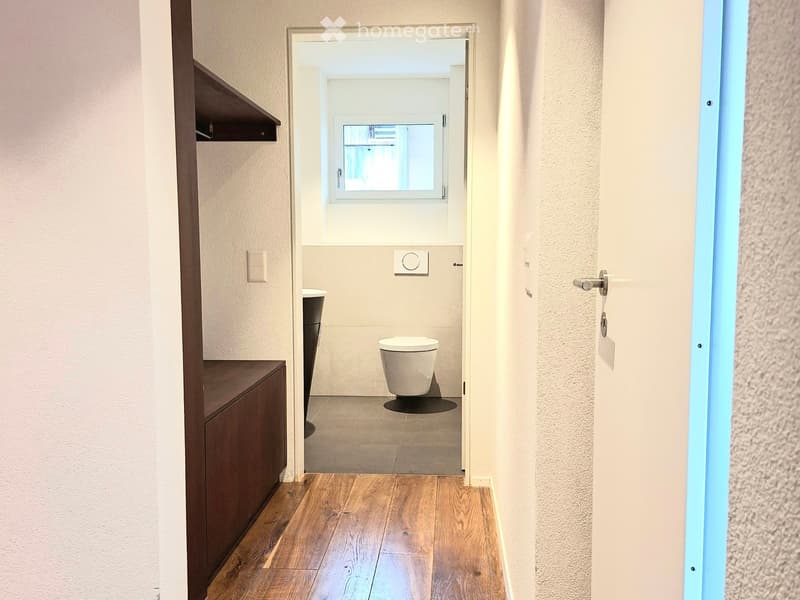 A hallway leading to a bathroom. There is a toilet and a sink. There is a white wall and a gray tiled floor. There is a window above the toilet.