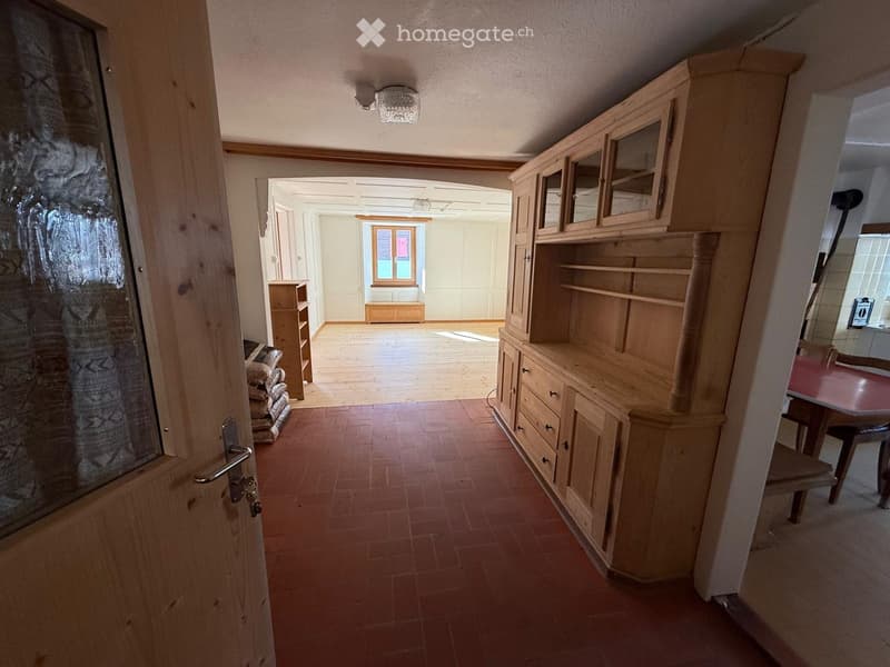 a wooden door leads into a hallway, a large wooden shelf is in the hallway, an arched door leads to a room with a window, a wooden table and chairs are to the right, the hallway has a tiled floor