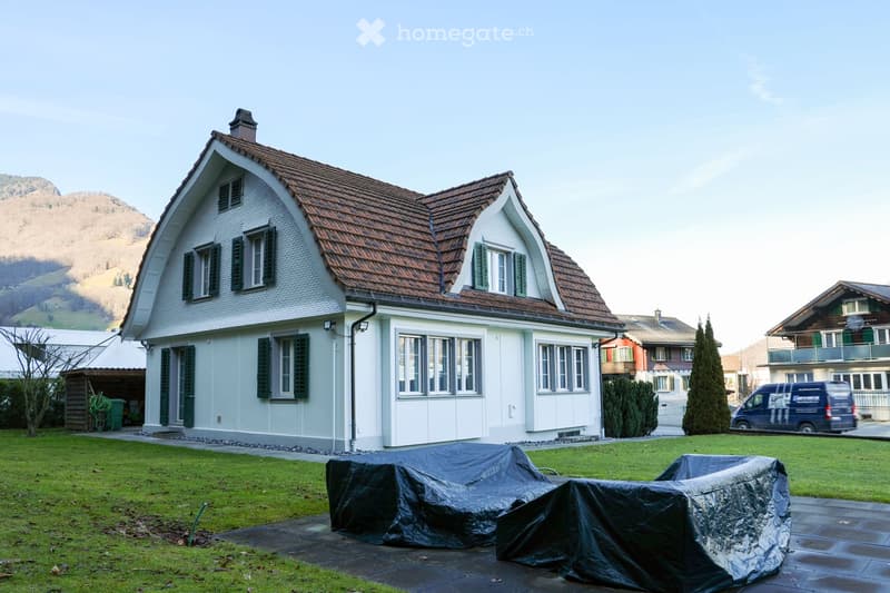two story house, white walls, brown roof, covered porch, green covered items in the front, garden, grassy lawn, mountains in the background