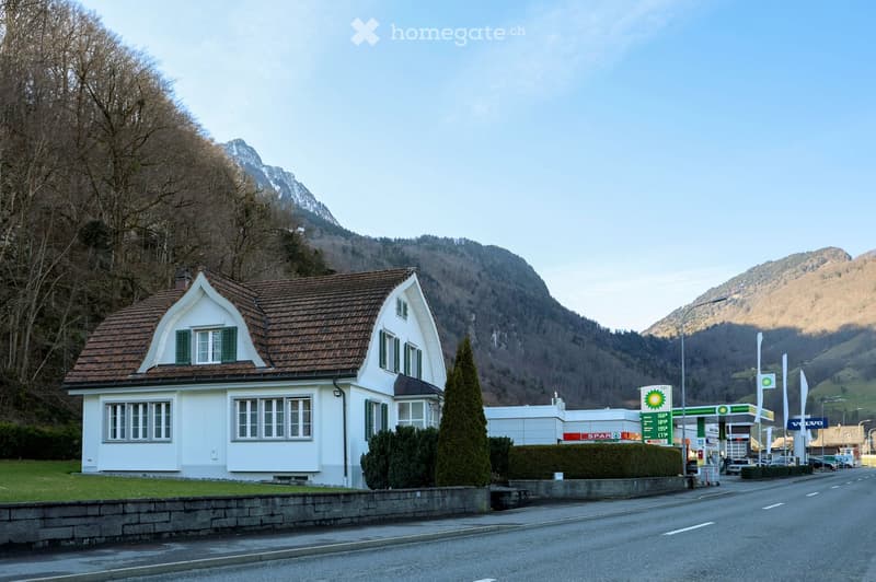 white house with brown roof, several windows, surrounded by greenery, mountains in the background