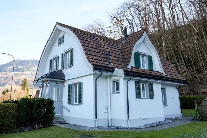 2 story white house, brown roof, green shutters, elevated ground floor, greenery in front, view of mountain